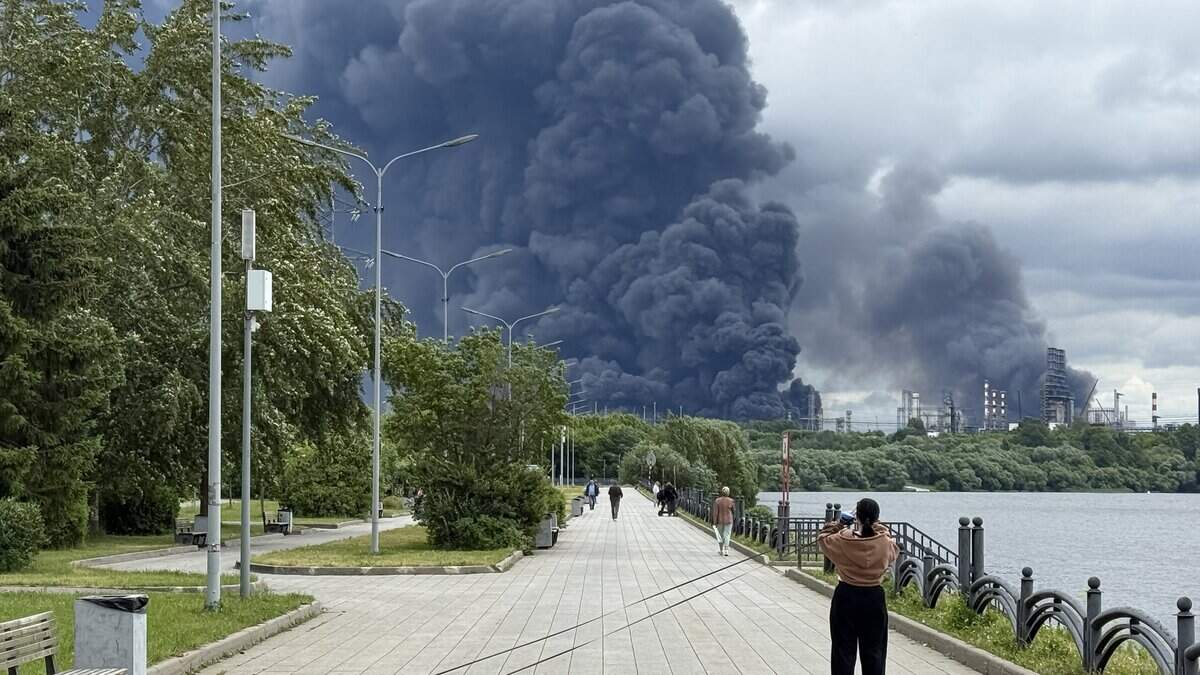 Personas caminan en un parque mientras una columna de humo negro se eleva desde la zona de la refinería de petróleo de Moscú del productor ruso Gazprom Neft, en las afueras del sureste de Moscú, el 18 de junio de 2026. Moscú enfrentaba un ataque “a gran escala” con drones procedentes de Ucrania, con varios aparatos que alcanzaron una refinería de petróleo, informó el alcalde de la ciudad, Serguéi Sobianin, a primera hora del 18 de junio de 2026. (