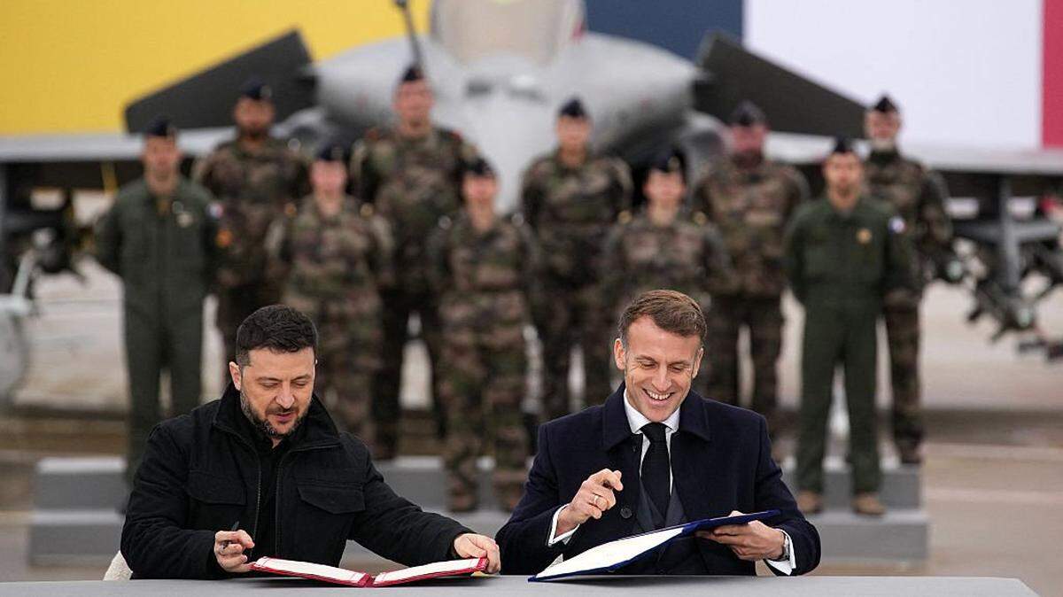 TOPSHOT - France's President Emmanuel Macron (R) and Ukraine's President Volodymyr Zelensky (L) sign an agreement at the Villacoublay air base, in Velizy-Villacoublay, near Paris on November 17, 2025. Macron and Zelensky signed an arms agreement at an airbase near Paris, which the Ukrainian president described as "historic" to "strengthen" Ukraine's combat aviation and air defense. (Photo by Christophe Ena / POOL / AFP) (Photo by CHRISTOPHE ENA/POOL/AFP via Getty Images)
