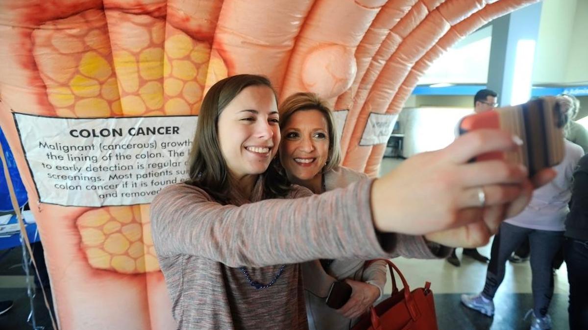 La sobreviviente de cáncer Susan Reese Burke (der.) con su hija Abbey mientras caminan a través de un colon enorme que muestra varios estadios de cáncer de colon.