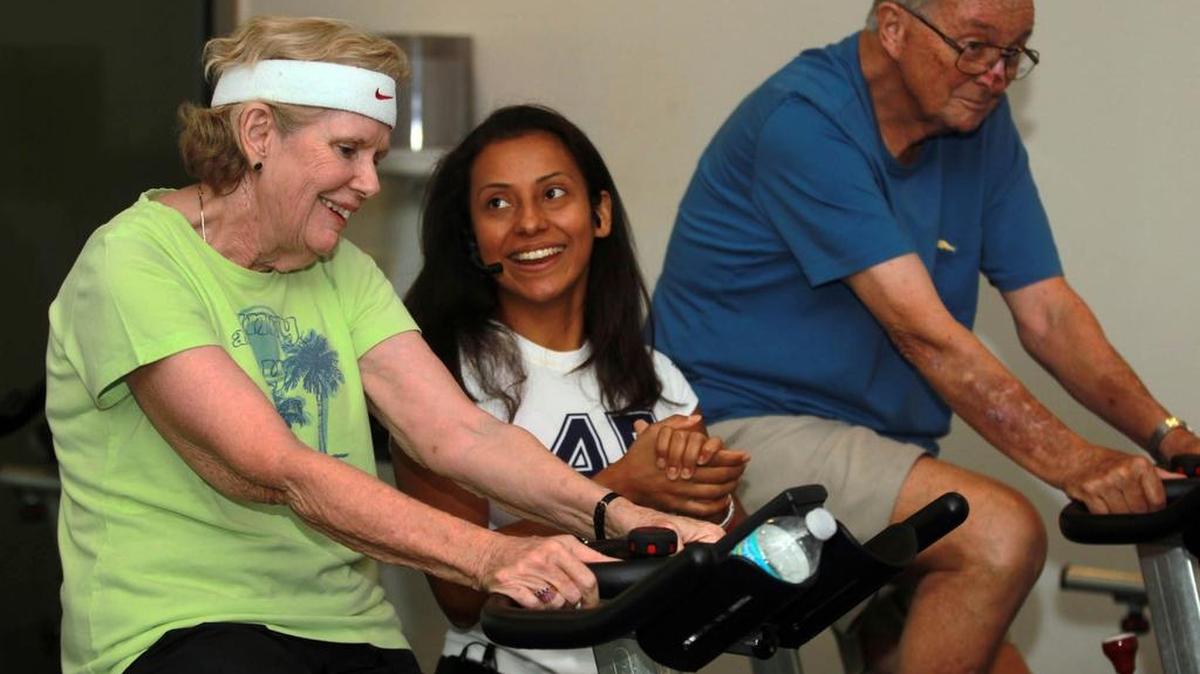 
Angela Alvarado, entrenadora de UHealth Wellness Center, ayuda a Claire Hackett, (izq) y a su marido, Bob Hackett, (dcha) en un rutina de ejercicios en bicicleta como parte de una clase para ayudar a pacientes de Parkinsons.

